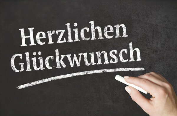 Eine Hand schreibt mit weißer Kreide die Worte 'Herzlichen Glückwunsch' auf eine schwarze Tafel.