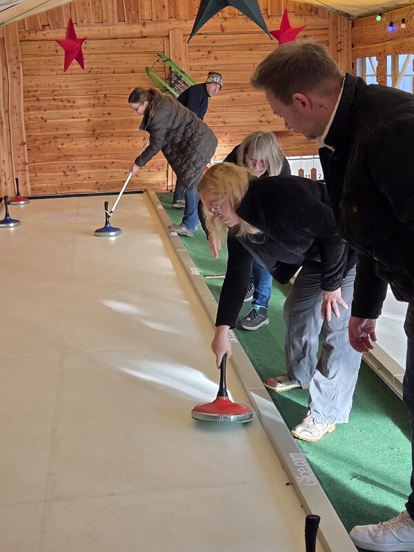 Mehrere Personen spielen Eisstockschießen in einer überdachten Halle aus Holz, eine Frau stößt gerade einen roten Eisstock an
