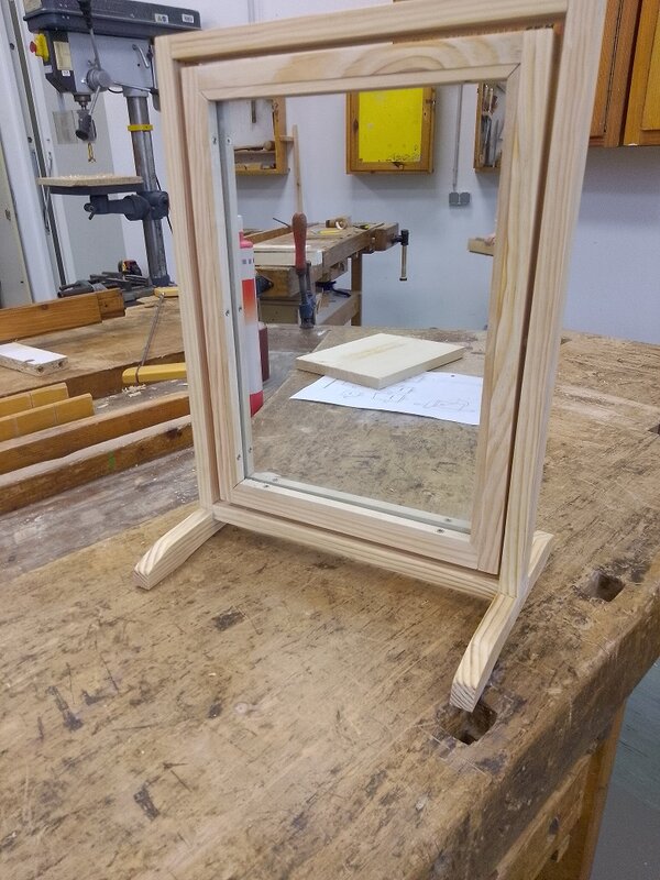Selbst gebauter, freistehender Spiegel mit hellem Holzrahmen und zwei Standfüßen auf einer Werkbank in einer Holzwerkstatt.