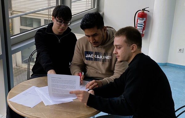 Drei Jugendliche mit Migrationshintergrund sitzen an einem kleinen, runden Holztisch im Gebäude der Euro-Schulen Leipzig. Vor ihnen liegen Formulare, die sie gemeinsam lesen und ausfüllen. Der Raum hat einen hellen Boden, weiße Wände und Metallstühle, im Hintergrund sieht man einen Feuerlöscher, der an der Wand befestigt ist. Die Szene vermittelt eine Gruppenarbeit und die aktive Teilnahme am Projekt KarriereStart III. 