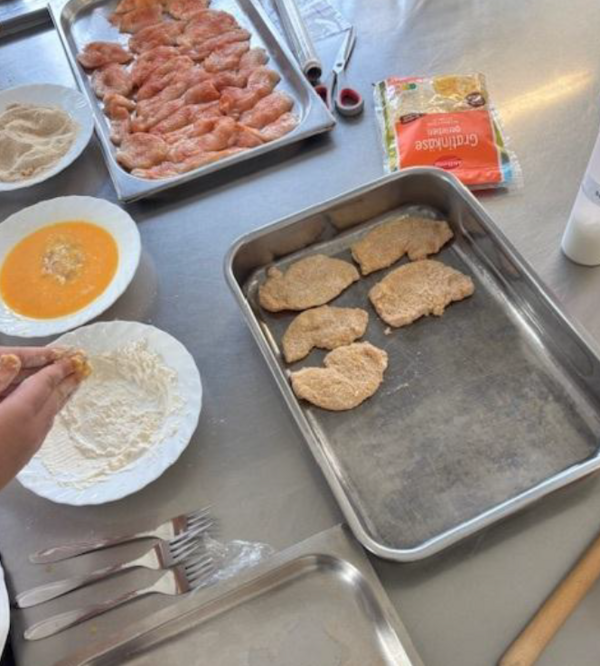 Vorbereitung von panierten Fleischstücken in einer Lehrküche. Auf einer Arbeitsfläche stehen Bleche mit gewürztem und bereits paniertem Fleisch sowie Teller mit Mehl, Ei und Paniermehl.
