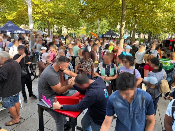 Belebte Szene auf dem Straßenfest im Park: Viele Besucherinnen und Besucher stehen an Ständen und in Gruppen zusammen, im Vordergrund zwei Männer an einem Tisch machen Armdrücken.