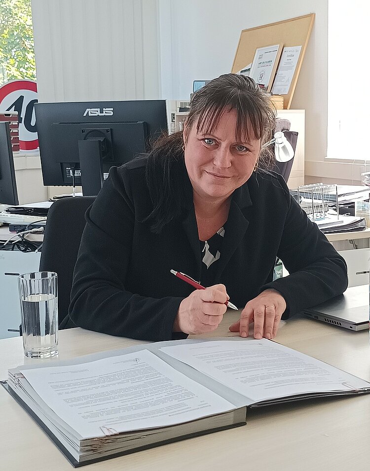 Unterzeichnung des Kooperationsvertrages im Projekt „Resilienz in der Kinder- und Jugendarbeit (Kita-Resilienz)“ Frau Doreen Saupe sitzt an einem Schreibtisch in einem Büro, hält einen Stift und unterschreibt ein Dokument. Neben ihr ein Glas Wasser, im Hintergrund Computer und Ordner.