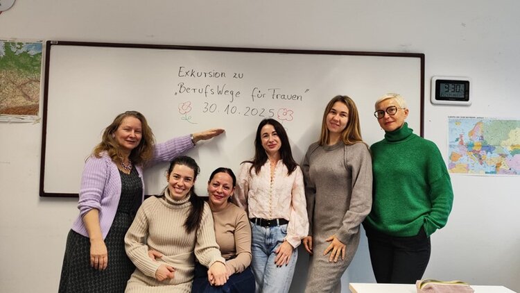 Sechs Frauen stehen vor einer weißen Tafel in einem Klassenraum. Auf der Tafel steht handschriftlich 'Exkursion zu "BerufsWege für Frauen"  mit einer kleinen Blume als Verzierung. Die Frauen tragen unterschiedliche Kleidung: eine trägt eine fliederfarbene Strickjacke über einem schwarzen Kleid, eine andere einen hellbeigen Rollkragenpullover, eine weitere eine beige Bluse, eine weitere eine graue Strickjacke, und die letzte eine grüne Strickjacke. An der Wand rechts neben der Tafel hängen eine analoge Uhr und eine bunte Landkarte. Die Szene wirkt freundlich und zeigt eine Gruppe von Teilnehmerinnen der Euro-Schulen Wiesbaden während einer Exkursion zum Verein BerufsWege für Frauen e.V.
