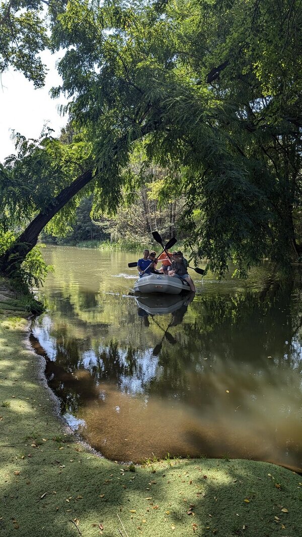 Ein Schlauchboot befindet sich im Schatten unter Bäumen auf einem kleinen Fluss. Die Besatzung hat zwei der vier Ruder in der Luft überkreuzt. Im Vordergrund ist das begrünte Ufer erkennbar.