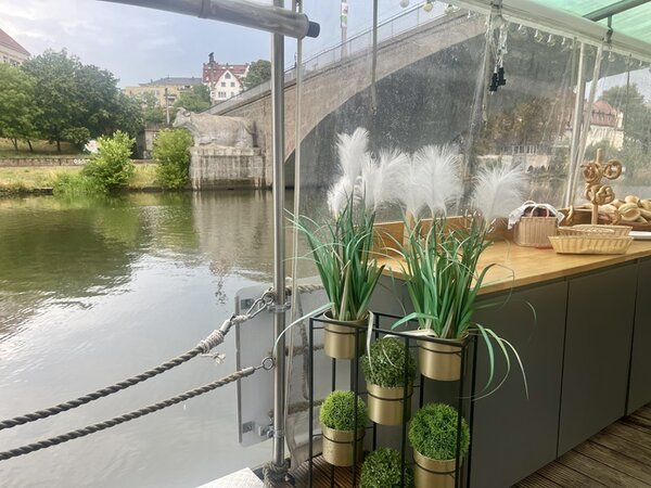 Blick von einem überdachten Boot oder Ponton auf einen Fluss mit steinernem Brückenbogen im Hintergrund. Im Vordergrund stehen dekorative Pflanzen in goldfarbenen Töpfen auf einer grauen Theke. Daneben liegen Brotkörbe und ein Tablett mit Gebäck. Am Ufer wachsen Bäume und Sträucher, dahinter sind Gebäude zu sehen.