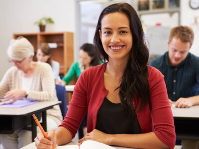 Eine lächelnde Frau mit langen dunklen Haaren und einem roten Cardigan sitzt an einem Tisch in einem Klassenzimmer. Sie hält einen Stift und schreibt in ein Notizbuch. Im Hintergrund sind mehrere Personen, die ebenfalls an Tischen arbeiten, darunter eine ältere Frau und ein Mann.