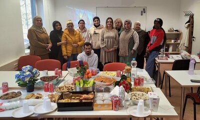 Eine größere Gruppe von Kursteilnehmenden posiert hinter einem reich gedeckten Buffet-Tisch im Klassenraum, in der Mitte sitzt ein Mann, im Hintergrund läuft per Beamer ein Weihnachtslied mit deutsch-englischem Text.