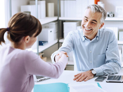 Eine junge Frau und ein älterer Mann sitzen an einem Tisch in einem Büro oder Beratungsraum. Die Frau, mit einem lila Pullover und einem Pferdeschwanz, reicht dem Mann lächelnd die Hand. Der Mann, mit graumeliertem Haar und einem hellblauen Hemd, schüttelt ihre Hand und lächelt ebenfalls. Auf dem Tisch liegen ein Laptop, ein Tablet, ein Smartphone und einige Dokumente. Im Hintergrund sind Regale mit Ordnern und Büromaterial zu sehen. Beide wirken freundlich und zufrieden, was auf eine positive und erfolgreiche Interaktion hindeutet.