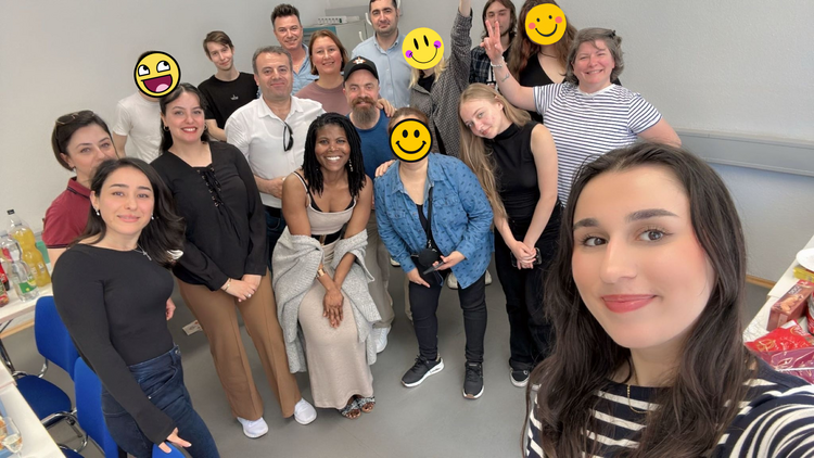 Gruppen-Selfie eines B2-Kurses in einem Klassenraum; mehrere Teilnehmende stehen eng zusammen und lächeln in die Kamera, einige Gesichter sind mit Smileys verdeckt.