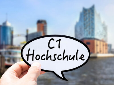 Eine Hand hält eine Sprechblase mit der Aufschrift 'C1 Hochschule' in den Vordergrund. Im Hintergrund ist die Skyline von Hamburg mit modernen Hochhäusern und dem Hafen zu sehen. Der Himmel ist klar und blau, und die Architektur der Gebäude ist deutlich erkennbar.