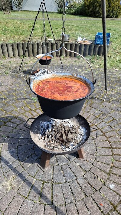 Ein großer schwarzer Kessel, der randvoll mit einer roten Suppe gefüllt ist, hängt über einem offenen Feuer im Freien. Darunter befindet sich ein Metallrost mit glühender Holzkohle. Im Hintergrund sind weitere Töpfe, Küchenutensilien, eine Wiese und Bäume zu sehen.