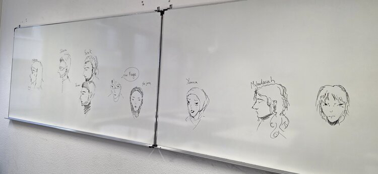 Ein Whiteboard auf dem mit schwarzem Edding verschiedene Köpfe und Gesichter gemalt sind.