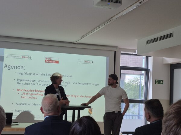 Zwei Personen sprechen bei einer Veranstaltung vor einer Agenda-Folie, während das Publikum im Raum zuhört.