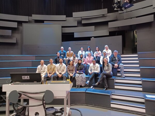 Eine Gruppe von Menschen sitzt im Publikumsbereich eines Fernsehstudios mit modernem, stufenförmigem Design. Ein Scheinwerfer ist auf sie gerichtet und leuchtet sie an. Die Personen sitzen in drei Reihen auf den Stufen. Im Vordergrund ist ein Tisch mit der Aufschrift "Discovery" und einem Monitor zu sehen.