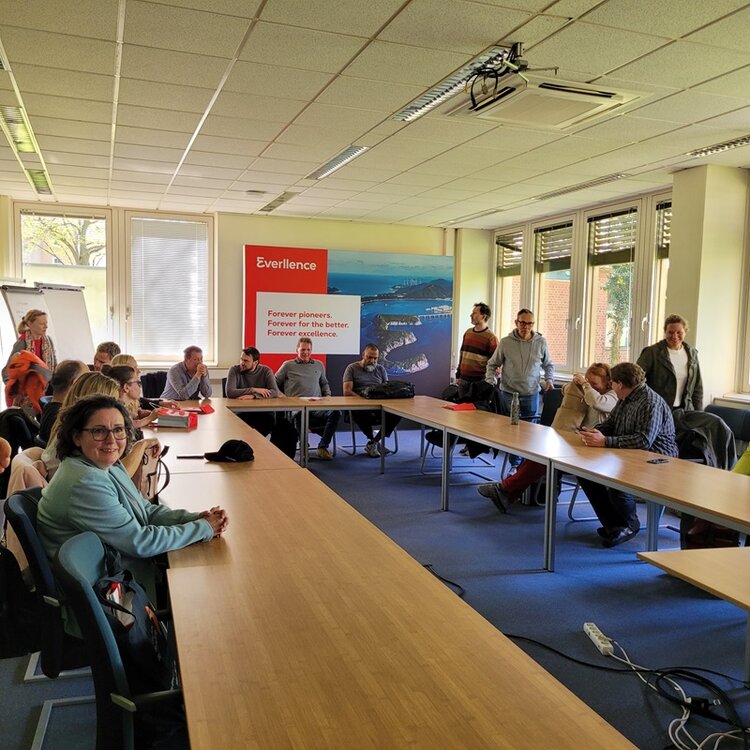 In einem Konferenzraum mit großen Fenstern sitzen und stehen mehrere Teilnehmende um zwei lange, rechteckige Tische, die zu einem U angeordnet sind. Im Hintergrund hängt ein großformatiges Wandbild mit dem roten Schriftzug 'Everience' und englischem Text. Die Personen sind überwiegend lässig gekleidet und blicken in verschiedene Richtungen. Die Decke ist mit Rasterplatten und Leuchtstoffröhren ausgestattet. Tageslicht fällt durch die Fenster, einige Jalousien sind halb geschlossen.