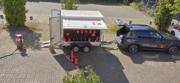 Ein dunkler Kombi mit geöffneter Heckklappe steht auf einem gepflasterten Parkplatz neben einem Anhänger. Der Anhänger ist seitlich geöffnet und enthält zahlreiche rote Feuerlöscher, die in Halterungen stehen. Einige Feuerlöscher stehen auf dem Boden vor dem Anhänger. Im Hintergrund sind Bäume und ein Zaun zu sehen. Die Szene wirkt wie eine Vorführung oder Wartung von Feuerlöschern.