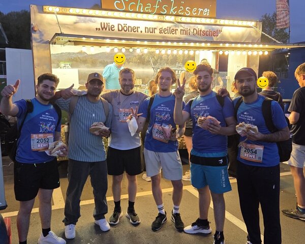 Die Teilnehmenden der Euro Akademie und der Euro-Schulen Jena posieren, gemeinsam mit Leiter Uwe Schöneck, in der Dämmerung und nach dem erfolgreich absolvierten Lauf, gemeinsam vor einem Schafskäse-Stand für ein Foto.