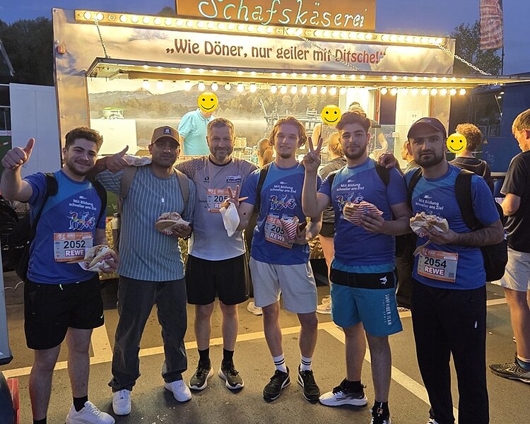 14. Jenaer Firmenlauf – Teamgeist und Begeisterung im Ernst-Abbe-Sportfeld Die Teilnehmenden der Euro Akademie und der Euro-Schulen Jena posieren, gemeinsam mit Leiter Uwe Schöneck, in der Dämmerung und nach dem erfolgreich absolvierten Lauf, gemeinsam vor einem Schafskäse-Stand für ein Foto.