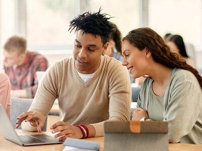 Zwei Personen sitzen an einem Tisch in einem Klassenzimmer oder einer Lernumgebung. Die Person links, ein Mann mit einem beigen Pullover, arbeitet an einem Laptop. Die Person rechts, eine Frau mit einem grünen T-Shirt, beobachtet und scheint sich zu unterhalten oder zuzuhören. Beide lächeln und wirken engagiert in ihrer Arbeit oder Diskussion. Im Hintergrund sind weitere Personen zu sehen, die ebenfalls an Tischen sitzen und arbeiten.