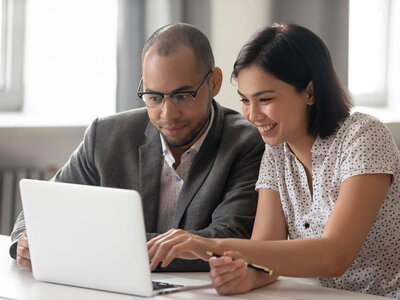 Ein Mann und eine Frau arbeiten gemeinsam an einem Laptop. Der Mann trägt eine Brille und einen grauen Anzug, während die Frau ein weißes, gepunktetes Oberteil trägt. Beide lächeln und scheinen sich auf den Bildschirm zu konzentrieren, während sie einen Stift in der Hand hält.