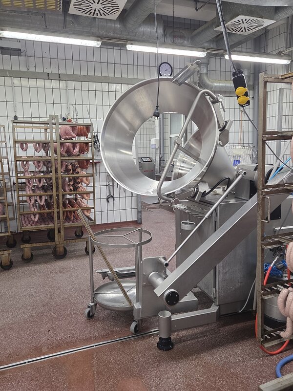 Große industrielle Edelstahlmaschine zur Verarbeitung von Wurstwaren in einer Produktionshalle; im Hintergrund stehen weitere Wagen mit Würsten.
