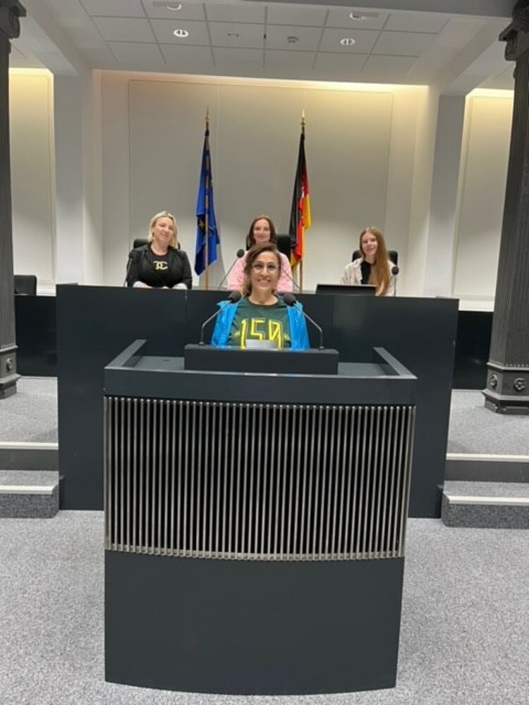 Politik hautnah erleben Im niedersächsischen Landtag steht eine Schülerin am Rednerpult. Dahinter sitzen drei weitere Schülerinnen an einem erhöhten Tisch, an dem ebenso Mikrofone platziert sind. An der rückwärtigen Wand stehen die Europaflagge und die deutsche Flagge nebeneinander. Der Boden ist mit Teppich ausgelegt, die Decke wird von zwei Säulen gestützt.