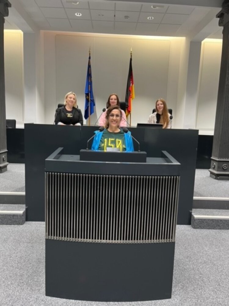 Im niedersächsischen Landtag steht eine Schülerin am Rednerpult. Dahinter sitzen drei weitere Schülerinnen an einem erhöhten Tisch, an dem ebenso Mikrofone platziert sind. An der rückwärtigen Wand stehen die Europaflagge und die deutsche Flagge nebeneinander. Der Boden ist mit Teppich ausgelegt, die Decke wird von zwei Säulen gestützt.