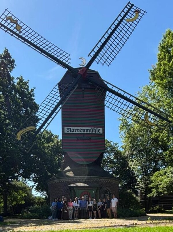 Gruppe von Menschen vor der historischen Narrenmühle, einer traditionellen Windmühle mit rot-grün gestreiftem Turm und goldenen Flügelenden, umgeben von hohem Grün und blauem Himmel.