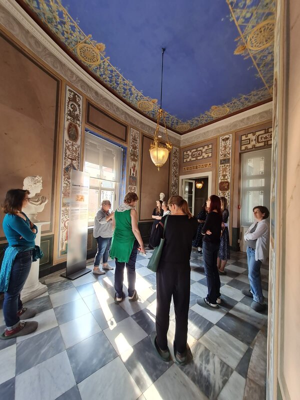 Eine Gruppe besichtigt einen Raum in einem Schloss. Der Boden ist aus Marmor und schwarz-weiß kariert, die Decke ist blau. Die Wände und die Decke sind mit Malereien verziert. Es hängt eine große historische Lampe von der Decke. 