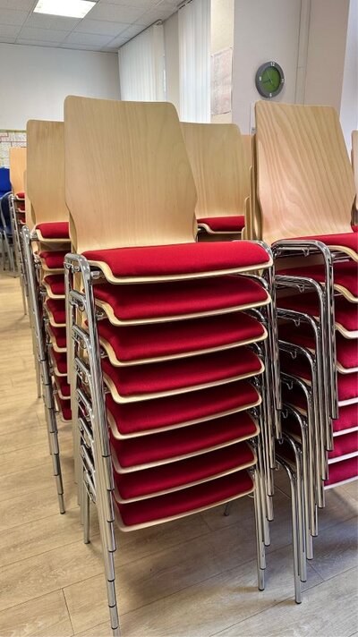 Ein Bild von mehreren gestapelten Stühlen mit Holzsitzflächen und roten Polstern in einem hellen Raum. Die Stühle haben Metallbeine und sind ordentlich aufeinandergestellt.
