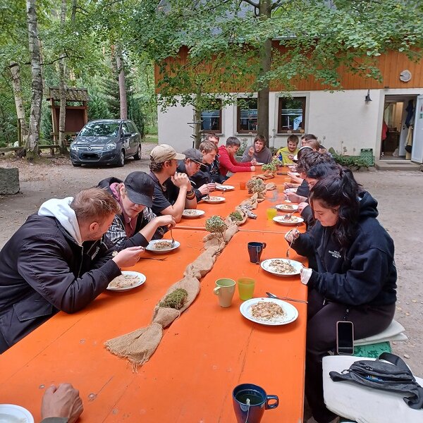 An einem langen Holztisch im Freien sitzt eine Gruppe Auszubildender beim Mittagessen. In der Mitte des Tisches liegen herbstliche Dekorationen. Im Hintergrund sind ein Haus, ein Auto und Bäume zu sehen.