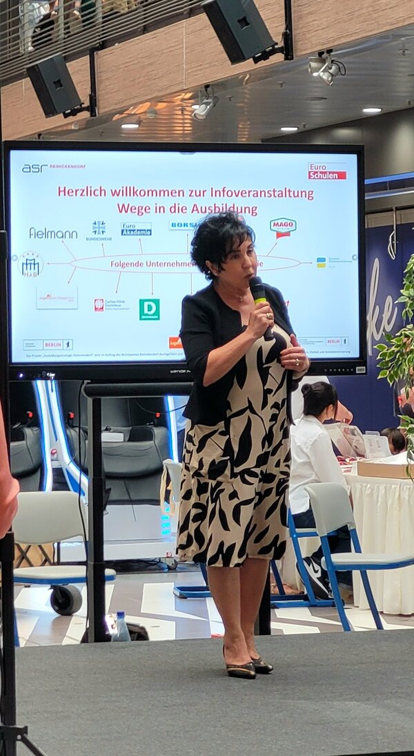 Die Bezirksbürgermeisterin Emine Demirbüken-Wegner begrüßt die Gäste der Veranstaltung, hinter ihr ein Bildschirm, au der alle beteiligten Unternehmen aufgeführt sind.