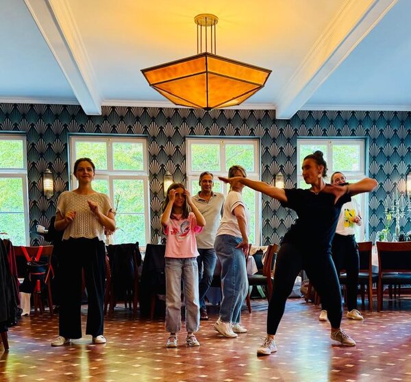Eine Gruppe von Menschen steht in einem Raum mit dekorativen Wandmustern und großen Fenstern. Einige Personen führen Bewegungen aus, während andere zuschauen. Der Raum wird von einer großen, auffälligen Deckenlampe warm beleuchtet.