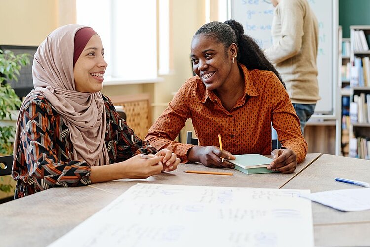MiA – Migrantinnen einfach stark im Alltag Zwei lächelnde Frauen sitzen an einem Tisch in einem hellen Raum, möglicherweise einer Bibliothek oder einem Klassenzimmer. Eine der Frauen trägt ein Kopftuch und hält die Hände der anderen Frau, während diese ein Notizbuch und einen Stift hält. Im Hintergrund sind Bücherregale und eine weitere Person zu sehen.