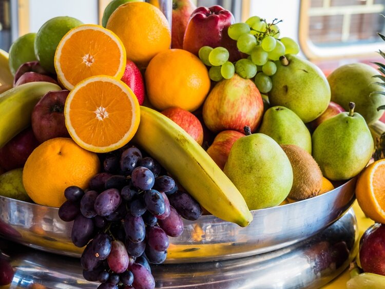 Verschiedene Früchte liegen in einer großen, glänzenden Metallschale. Im Vordergrund sind eine aufgeschnittene Orange, eine Banane und dunkle Weintrauben zu sehen. Dahinter befinden sich grüne Birnen, rote Äpfel, grüne Weintrauben, eine Kiwi und weitere Zitrusfrüchte. Die Farben der Früchte sind kräftig und frisch. Im Hintergrund ist ein unscharfer Innenraum mit Fenstern erkennbar.