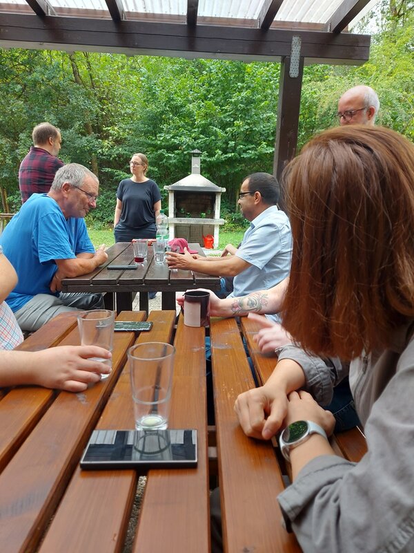 Mehrere Mitarbeitende sitzen gemeinsam an Holztischen unter einer überdachten Terrasse. Sie unterhalten sich bei Getränken; im Hintergrund stehen ein gemauerter Grill und dicht bewachsene Bäume.