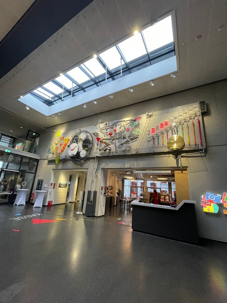 Innenbereich der Phänomenta in Lüdenscheid während der Veranstaltung der Euro-Schulen Märkischer Kreis: Eine moderne, hohe Eingangshalle mit großem Oberlicht, glatten dunklen Fußböden und einer großformatigen interaktiven Wandinstallation. Der Raum dient als Empfangs- und Orientierungspunkt für das Job-Speed-Dating. Farbenfrohe Markierungen auf dem Boden weisen Besucherinnen und Besucher auf verschiedene Bereiche hin. Links befindet sich ein Eingang zu weiteren Räumen, rechts ein Empfangstresen. Die Atmosphäre wirkt offen, hell und einladend.