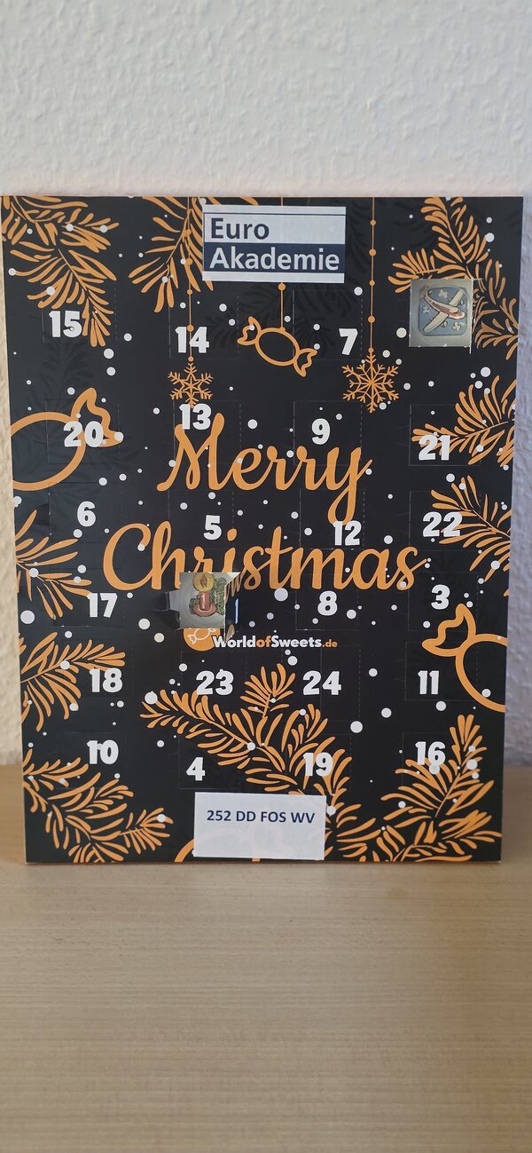 Schwarzer Adventskalender mit orange-goldenen Tannenzweigen und der Aufschrift „Euro Akademie – Merry Christmas“, die Türchen sind mit weißen Zahlen beschriftet, einige sind geöffnet und geben kleine Bildchen frei; unten klebt ein Etikett mit der Gruppenbezeichnung „252 DD FOS WV“.
