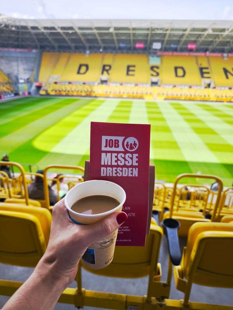 Im Vordergrund hält eine Hand einen Pappbecher mit Kaffee und einen roten Flyer mit weißem Aufdruck. Im Hintergrund ist ein Fußballstadion mit gelben Sitzreihen und dem Schriftzug 'DRESDEN' auf der Tribüne zu sehen. Das Spielfeld zeigt ein Muster aus hellen und dunklen Rasenstreifen. Die Szene ist von den oberen Sitzreihen aus fotografiert.
