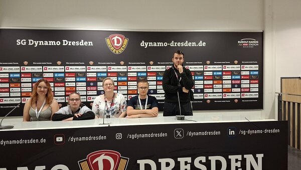 Die Gruppe sitzt am Tisch im Presseraum des Fußballvereins SG Dynamo Dresden, im Hintergrund die typische Sponsorenwand – ein authentischer Moment wie bei einer echten Pressekonferenz.