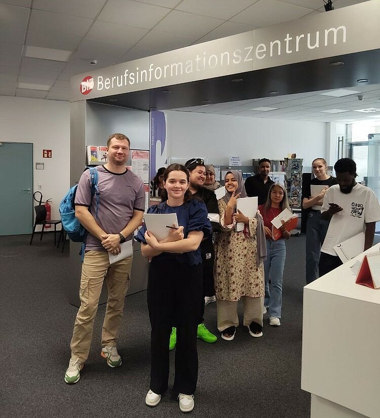 Gruppe IK-359 zu Besuch im BIZ Eine Gruppe von Teilnehmenden eines Integrationskurses steht lächelnd vor dem Eingang des Berufsinformationszentrums. Sie halten Informationsmaterialien in den Händen und wirken motiviert.