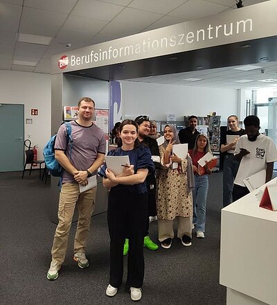 Gruppe IK-359 zu Besuch im BIZ Eine Gruppe von Teilnehmenden eines Integrationskurses steht lächelnd vor dem Eingang des Berufsinformationszentrums. Sie halten Informationsmaterialien in den Händen und wirken motiviert.