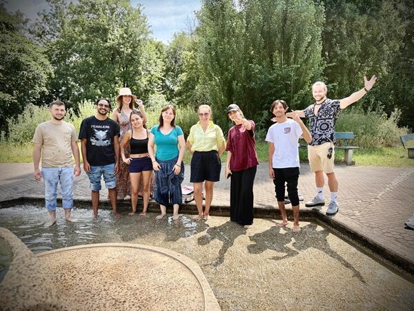 Die Gruppe der Teilnehmenden steht barfuß in einem flachen Wasserbecken im Freien. Die Personen sind sommerlich gekleidet, einige tragen kurze Hosen, T-Shirts oder Blusen, eine Person trägt einen Hut. Im Hintergrund sind Bäume, Sträucher und zwei leere Parkbänke zu sehen. Die Stimmung wirkt entspannt, einige Personen lächeln oder gestikulieren. Das Licht ist hell, der Himmel teilweise bewölkt.