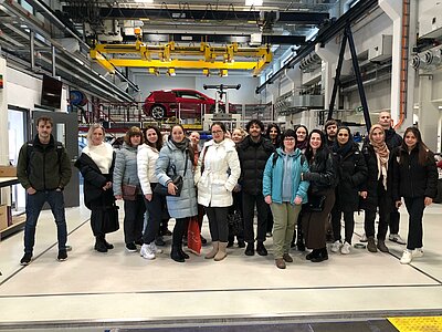 Sonniger Exkursionstag der Euro-Schulen Dresden: Spannende Einblicke in Wissenschaft und Kultur Eine große Gruppe von Personen steht in einer Werkshalle oder einem technischen Labor mit einem roten Auto im Hintergrund. Sie tragen Winterjacken und posieren für ein Gruppenfoto.