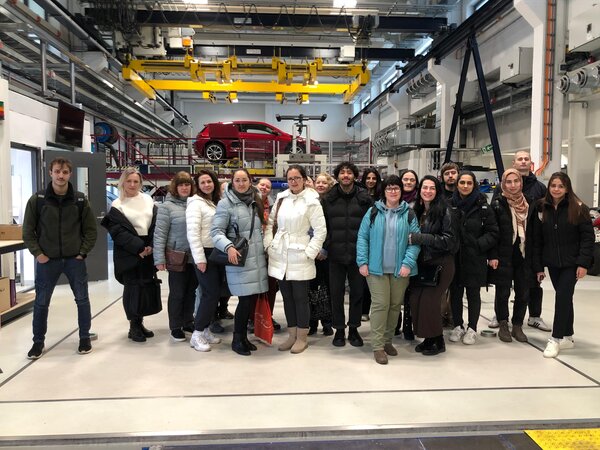 Eine große Gruppe von Personen steht in einer Werkshalle oder einem technischen Labor mit einem roten Auto im Hintergrund. Sie tragen Winterjacken und posieren für ein Gruppenfoto.