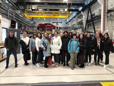 Eine große Gruppe von Personen steht in einer Werkshalle oder einem technischen Labor mit einem roten Auto im Hintergrund. Sie tragen Winterjacken und posieren für ein Gruppenfoto.