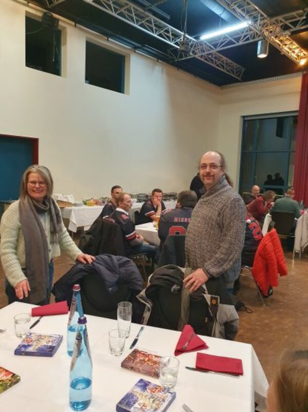 In einem hellen Saal stehen eine Frau und ein Mann lächelnd an einem festlich gedeckten Tisch mit Tellern, Gläsern und Wasserflasche. Im Hintergrund sitzen weitere Gäste in legerer Kleidung und Trikots zusammen und unterhalten sich.