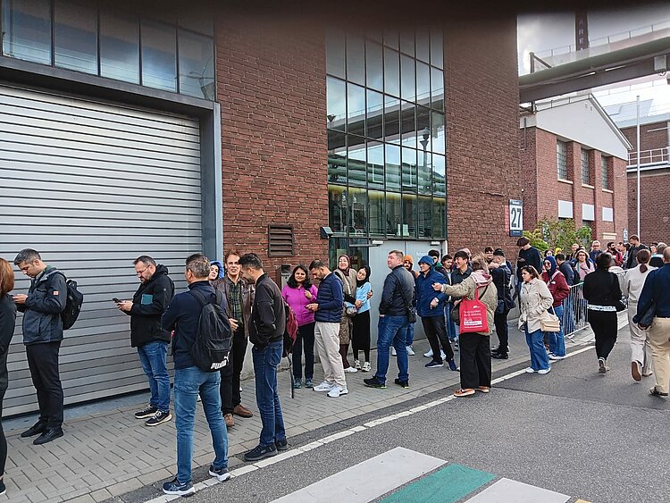 Blick auf die Schlange am Einlass vor der Messehalle aus braunen Backsteinen.