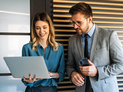 Eine junge Frau und ein junger Mann stehen in einem modernen Büro und arbeiten zusammen. Die Frau, mit langen blonden Haaren und einem blauen Blazer, hält lächelnd einen offenen Laptop in den Händen und schaut konzentriert auf den Bildschirm. Der Mann, mit Brille, Bart und einem grauen Anzug, hält ein Smartphone in der Hand und schaut ebenfalls auf den Laptop-Bildschirm der Frau. Beide wirken engagiert und kooperativ. Im Hintergrund sind große Fenster und eine moderne Büroausstattung zu sehen.